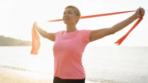 Mujer con camiseta rosa y los brazos extendidos, sosteniendo una banda elástica roja junto al mar.