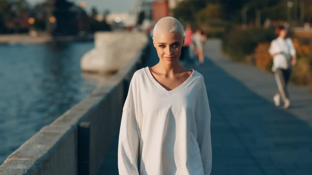 Mujer con cabeza rapada caminando al aire libre junto al río, con jersey blanco